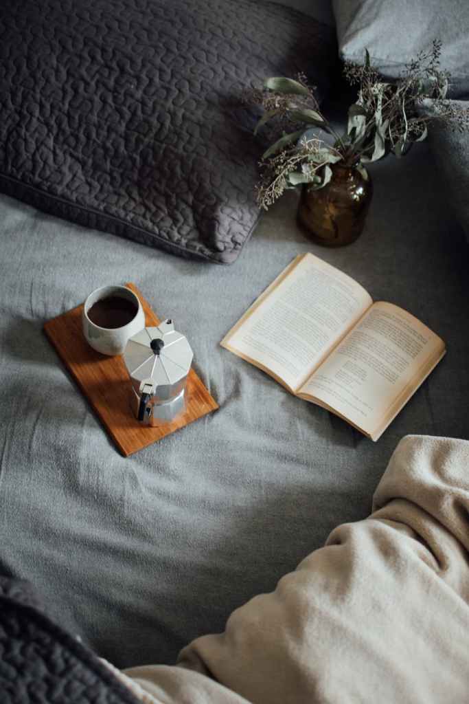 Photo of an open book sitting on a bed with a coffee tray and plant next to it.