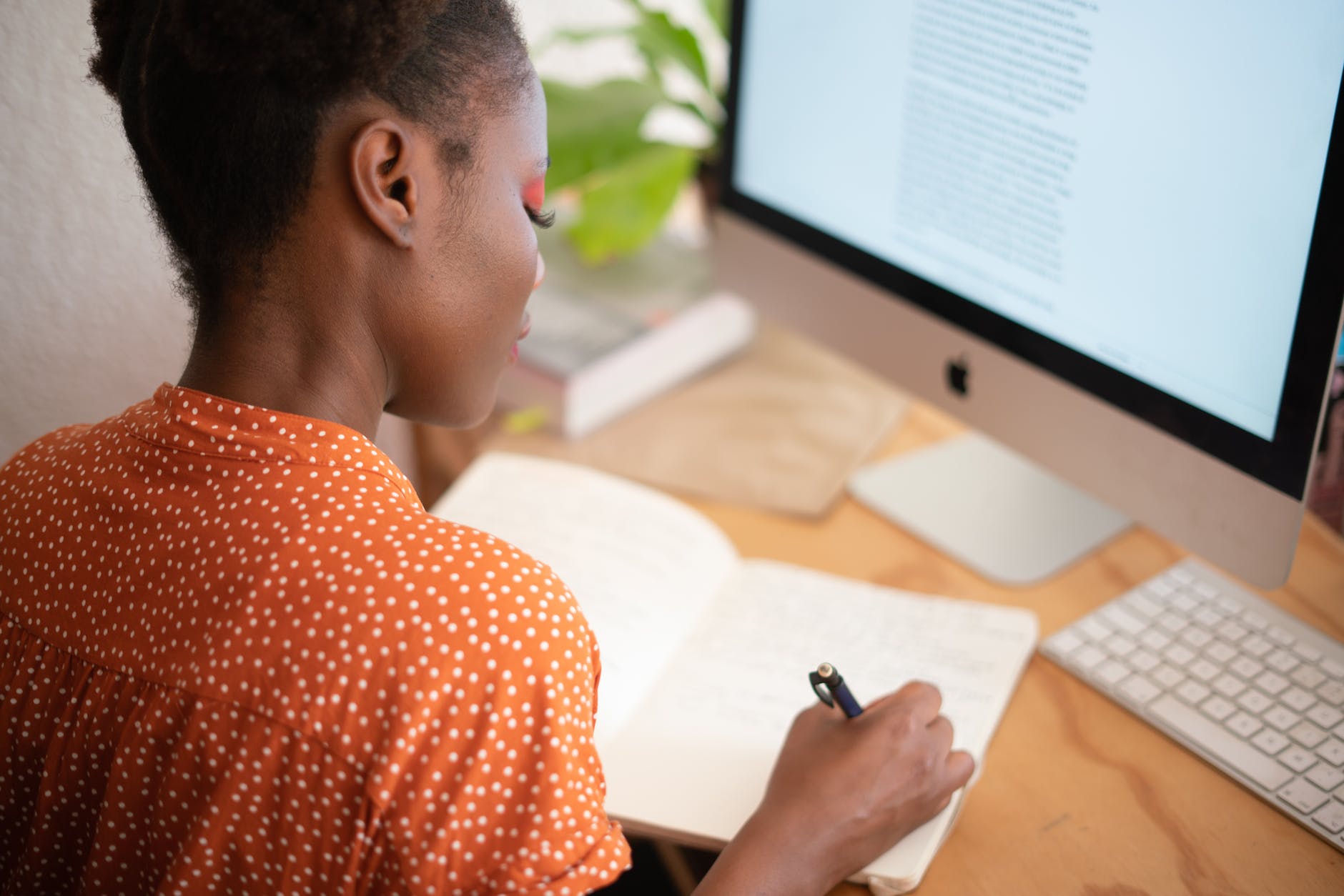 Image of a Black woman sitting in front of a desktop computer while writing in her notebook.