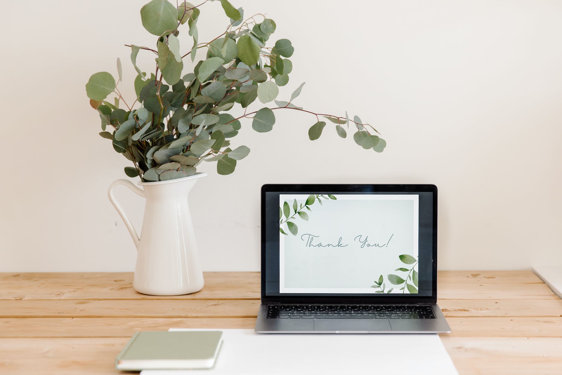 Image of a vase with green tables atop a light colored wood desk. On the desk is a laptop and a couple books. The laptop has an image pulled up that says "thank you."